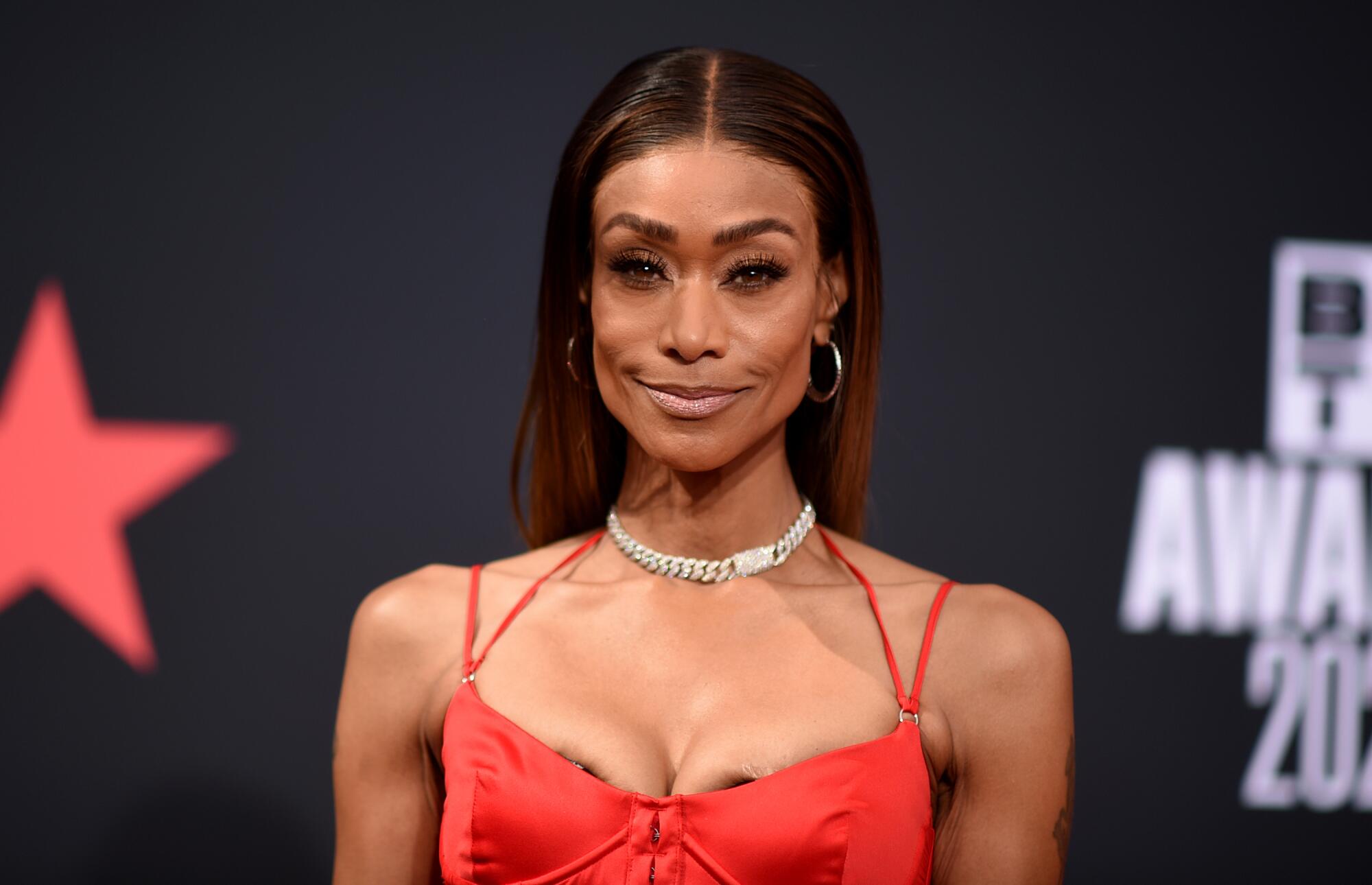 A headshot of Tami Roman standing in front of a black backdrop wearing a red dress and necklace.