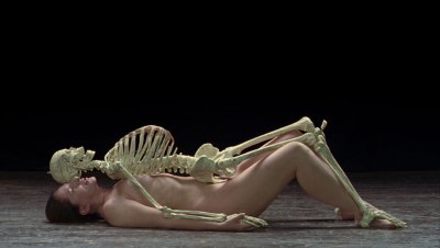 A nude white woman lies under a skeleton model.